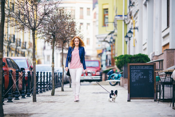young redhaired Caucasian woman walking along European street with small Chihuahua breed dog of two...