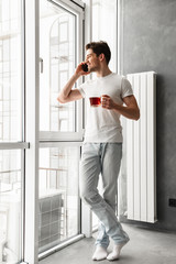 Fototapeta na wymiar Full length photo of joyful brunette man in casual wear speaking on mobile phone, and looking out the window while holding glass with tea