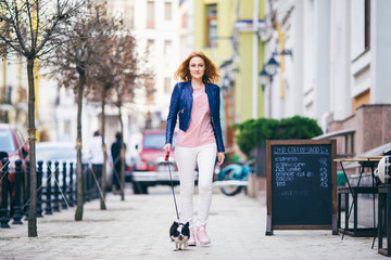 young redhaired Caucasian woman walking along European street with small Chihuahua breed dog of two...
