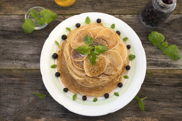 Pancakes are located in a plate on an old wooden table decorated with berries and mint leaves. Pancakes are watered with syrup.