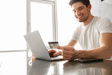 Photo of joyous adult man 30s in casual clothing holding credit card, while buying online using laptop in modern apartment