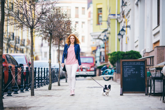 young redhaired Caucasian woman walking along European street with small Chihuahua breed dog of two colors on leash. Cloudy, warm autumn spring weather. Girl Dressed in leather jacket and pink shoes