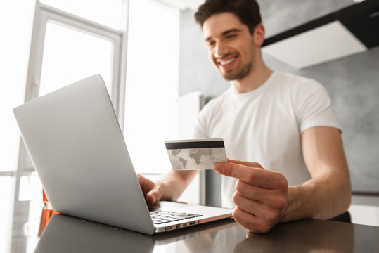 Photo of successful man 30s in casual clothing holding credit card, and doing payment transaction on laptop in modern apartment