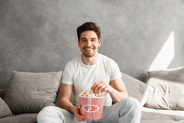 Cheerful bachelor sitting on sofa in gray apartment and laughing, while watching tv with pop corn...