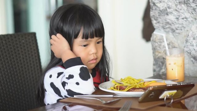 Asian children cute or kid girl smile with happy eating food with iced tea for breakfast or lunch and watch cartoon on smartphone because nomophobia or attention deficit hyperactivity disorder ADHD