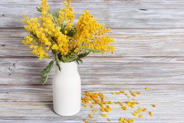 A bouquet of mimosa flowers in a white ceramic vase on wooden table. Yellow acacia in the vase is still life. Selective focus.
