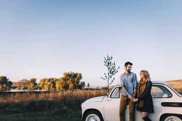 Young beautiful couple hugging each other, sitting on a small white car in beautiful evening light. Stylish guy with a beard and blond girl laughing