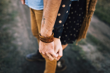 handsome guy with a beard in a denim shirt hugs and holds the hand of a beautiful girl in a blue dress and yellow scarf in a field at sunset. close-up hands together