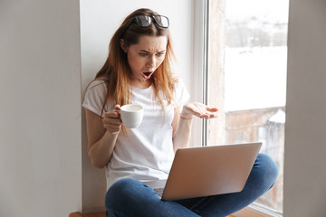 Shocked woman in t-shirt and eyeglasses sitting on windowsill