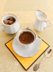 White porcelain cup of coffee, a sugar bowl with brown sugar, a milkman and a silver teaspoon on a table covered with silk tablecloth