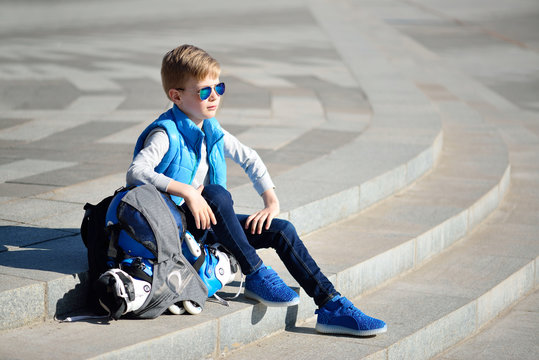 Young boy sitting near grey wall with inline roller skates and all protection at outdoor skate park