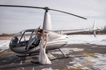 Beautiful girl in a helicopter. Bride. Wedding dress, wedding make-up and hairstyle. Fur coat. Looking sideways. Gray helicopter. Winter.