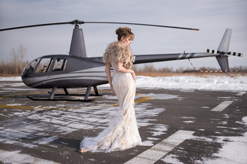 Beautiful girl in a helicopter. Bride. Wedding dress, wedding make-up and hairstyle. Fur coat. Look down. Standing with your back. Gray helicopter. Winter.