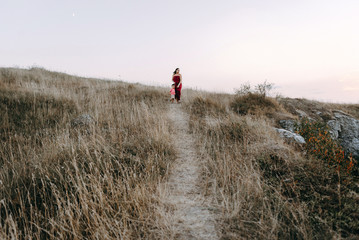 beautiful young mother in a long red dress easy walks with her young son. Summer, sunset, high yellow grass, mountains. He is on the hands, holding the hand