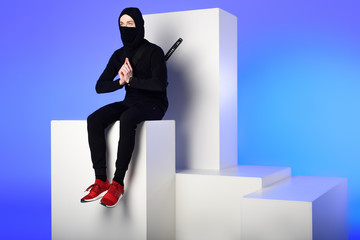 ninja in black clothing with katana behind sitting on white block isolated on blue