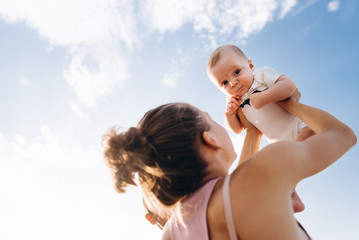 Mother holding her cute little toddler boy. Adorable child looking to camera in mother’s hands on sunny summer day on blue sky background