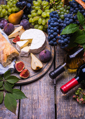 Red wine in a bottle and white wine bottle, cork, bottle screw and a set of products - cheese, grapes, nuts, pears, plums, flowers, figs, white bread, baguette on a wooden board, background