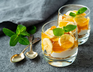 Healthy layered dessert with yogurt, banana, mango jam, cookie in glass on gray stone background, side view.