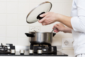 woman is cooking soup in kitchen. housewife prepares food at home. caucasian woman holds lid from saucepan in her hand and stirs soup in saucepan.