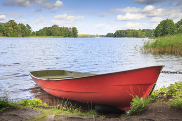 Red boat on shore on background of picturesque landscape of lake and green nature around in bright sunny summer day. Blue sky with white clouds over the lake.