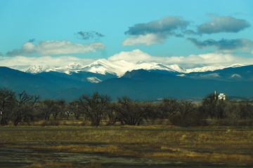 Snow covered rocky mountains rise high above a tree covered farmland 