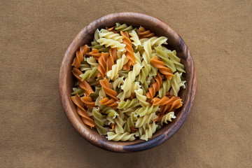 Multicolor spiral macaroni pasta in a wooden bowl on a brown rustic texture background, in the center close-up from the top.