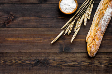 Fresh bread concept. Crispy french baguette near ears of wheat and bowl with flour on dark wooden background top view copy space