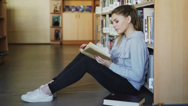 Young beautiful caucasian female student in casual clothing is sitting on floor in big spacious library near bookcase reading interesting big book seriously