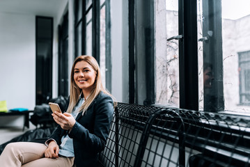 Portrait of a smiling blonde businesswoman using phone while sitting near the window.