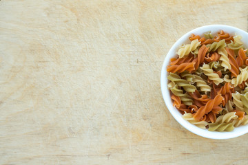 Multicolor spiral macaroni pasta in a white bowl on a wooden table textured background with a side. Close-up with the top. With space for text.