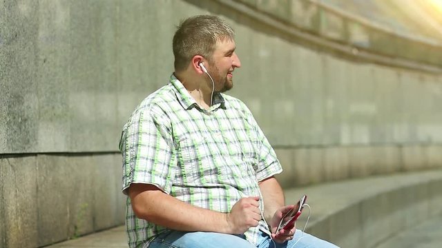 Man listens to music and moves. Businessman sits on the step and listens to music on his smartphone. Fat guy with red mobile phone