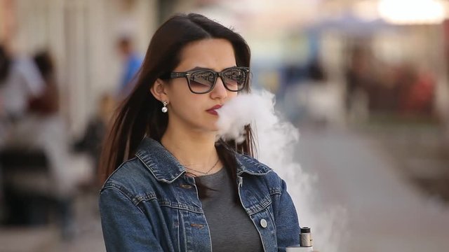 Portrait of an elegant young woman with long loose hair and sunglasses smoking an electronic cigarette and breathing out white fume in spring