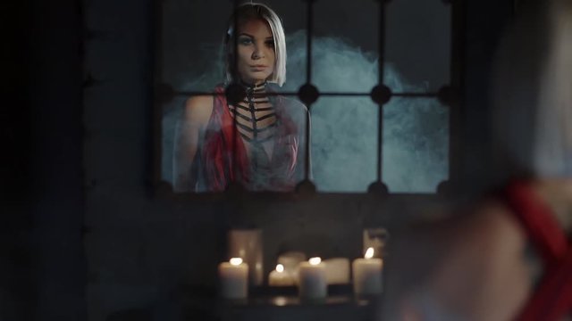 Mysterious blonde woman is looking in a mirror in dark hall in castle. She is coming to burning candles, moving through smoke