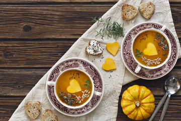 Close-up photo of two plates with fresh homemade pumpkin cream soup with seeds and heart shape toasts on vintage wooden background. Top view.