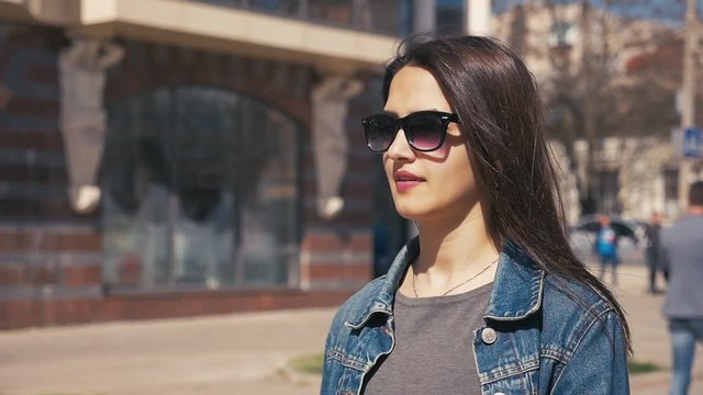 A splendid profile of a beautiful young woman in sunglasses walking along a city street in windy weather on a sunny day in slow motion