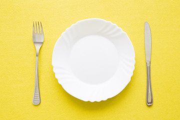 Clean, empty, white plate, fork and knife with yellow tablecloth on a table. Cutlery concept. Flat lay. Top view.