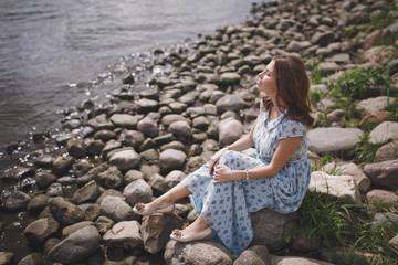 A beautiful young brunette woman with long hair in a long blue dress sits by the lake or river on a warm summer day.