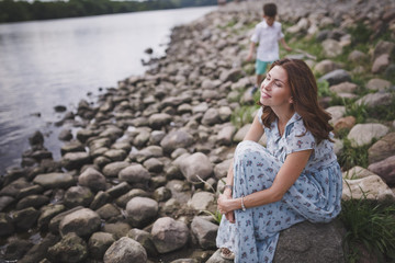 A beautiful young smiling brunette woman with long hair in a long blue dress sits on the shore of a lake or river. On the back of the blurry terms of boy walking.