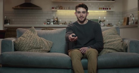 Young, attractive man sits down on a couch and turns on the television using a remote control. - Powered by Adobe