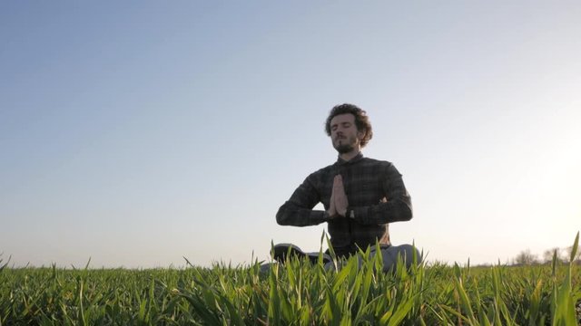 Young man in casual wear relaxing in green field after work, meditation of male against sunset or sunrise 