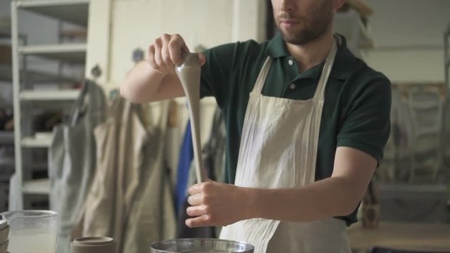 Portrait of young male ceramist, who is preparing clay fto work in his bright studio. Man in grey polo t-shirt and dickey is standing at the table, putting piece of grey clay into steel bowl with