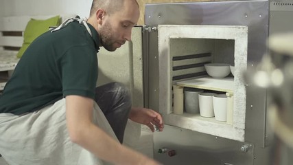 In the pottery workshop man puts clay pots for firing in the oven. Bearded craftsman in an apron places a billet in hot stove to create ceramic dishes.