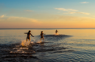 Three people run from shore to water at sunset