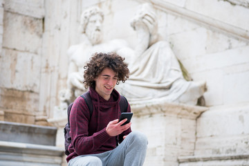Fototapeta na wymiar Handsome young man with curly hair in tracksuit using his mobile phone in front of Nile God statue in Rome
