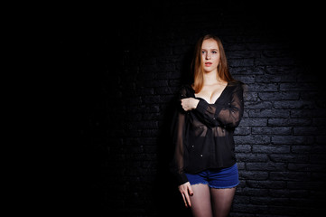 Studio shot of brunette girl in black blouse with bra and shorts against black brick wall.