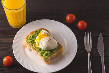 Breakfast. Toast with smashed avocado and poached egg on white plate with cherry tomatoes,  fresh orange juice on brown wooden background