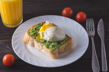 Breakfast. Toast with smashed avocado and poached egg on white plate with cherry tomatoes,  fresh orange juice on brown wooden background. Close-up