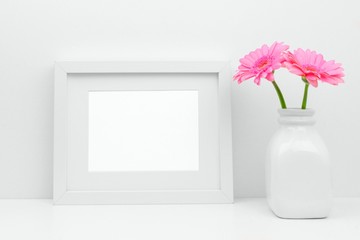 Mock up white frame and pink daisy flowers in vase on a shelf or desk. White color scheme. Landscape frame orientation.