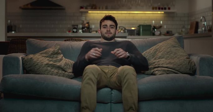 Man walks into the living room and sits on a couch to watch tv