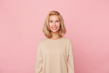 Indoor shot of young blonde female, feels satisfied and joyful, standing over pink wall and smiles. Isolated picture of glad woman in good mood